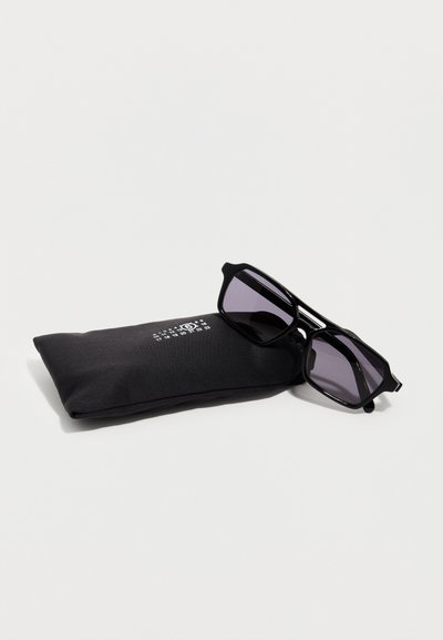 Γυαλιά ηλίου με μαύρο τετράγωνο πλαίσιο και σκουρόχρωμους φακούς, ακουμπισμένα πάνω σε μαύρη υφασμάτινη θήκη με λευκούς αριθμούς και κυκλικό λογότυπο.