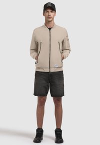 Beige bomberjakke med lynlås foran, to sidelommer og ribbede manchetter; parret med mørke shorts og sorte høj-top sneakers.