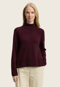 Maglione a collo alto bordeaux realizzato in morbido tessuto a maglia, con polsini e orlo a coste, abbinato a pantaloni chiari.