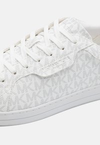 Baskets blanches avec un tissu texturé, motif de logo circulaire et lacets plats. Présentent une semelle blanche et un branding discret sur le côté.