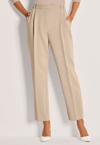 Vrouw in beige hoge taille plooibroek met zijzakken en witte puntige hoge hakken, staande tegen een effen achtergrond.