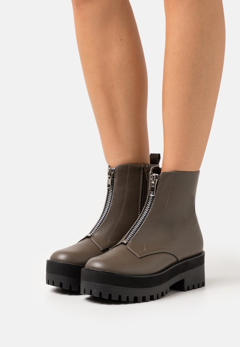 Bottes taupe montant à la cheville avec semelles crantées noires épaisses et fermetures éclair argentées à l'avant, portées sur jambes nues sur un fond uni.