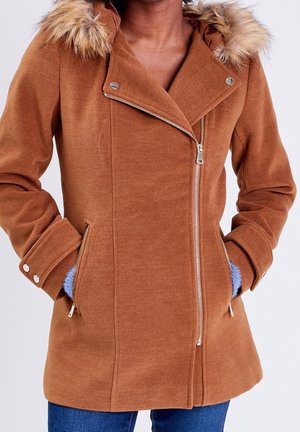 Manteau en laine marron avec fermeture éclair argentée asymétrique, col bordé de fourrure, poignets à boutons et mains dans les poches zippées sur un jean bleu.