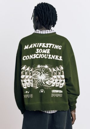 Osoba o ciemnej karnacji i dredach, ubrana w oliwkowozieloną bluzę z białym napisem i wzorem graficznym na plecach, stojąca na tle białego tła.