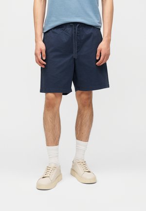 Hombre con pantalones cortos azul marino con cordón, calcetines blancos hasta el tobillo, zapatillas beige y una camiseta azul claro recortada a la altura de la cintura.