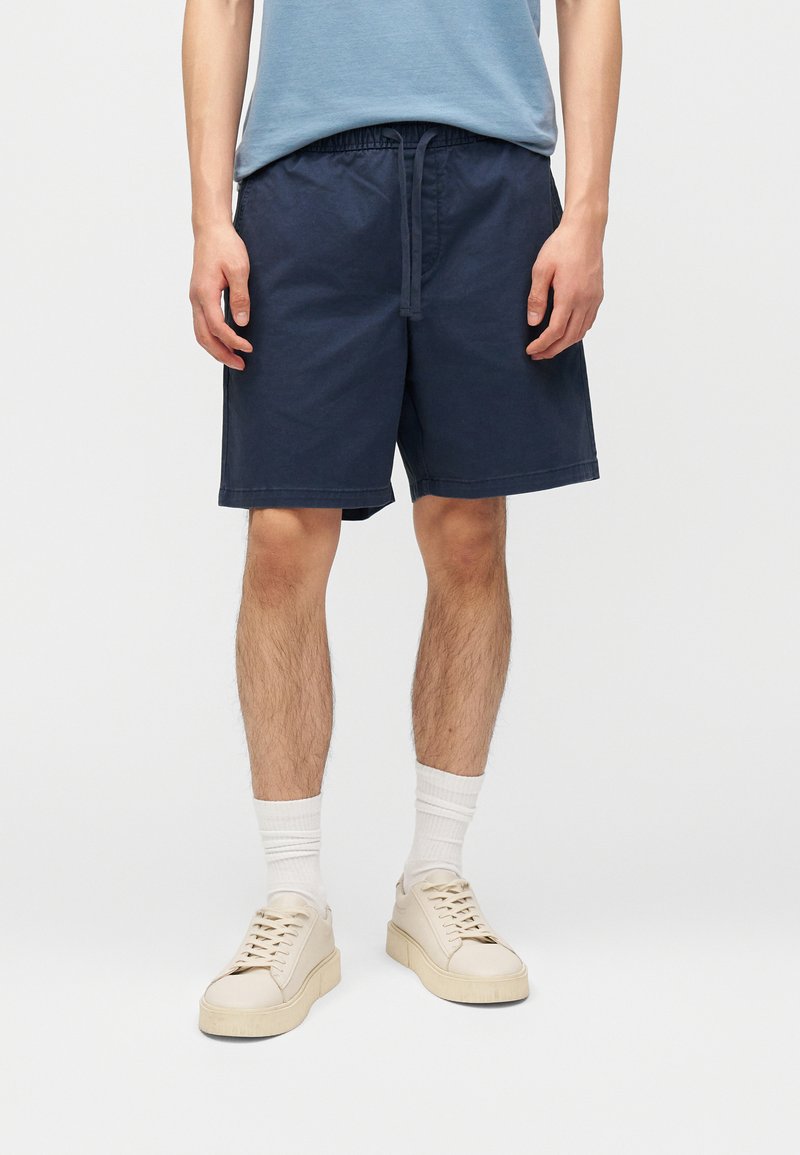 Άνδρας που φοράει ναυτικές βερμούδες με κορδόνι, λευκές κάλτσες μέχρι τον αστράγαλο, μπεζ αθλητικά παπούτσια και ανοιχτό μπλε μπλουζάκι κομμένο στη μέση.