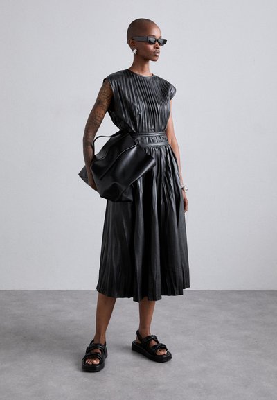 Schwarzes Lederkleid mit vertikalen Falten, tailliertem Bund und fließendem Rock. Kombiniert mit einer großen schwarzen Tasche und klobigen schwarzen Sandalen.