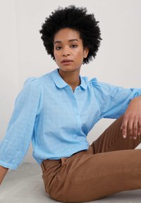 Svetlo modra srajca s gumbi in teksturiranim vzorcem ter napihnjenimi rokavi, v kombinaciji z rjavimi hlačami. Srajca ima ovratnik in sprednje gumbe.