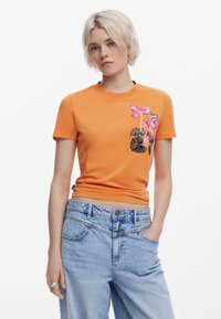 T-shirt in cotone arancione con ricami floreali rosa e marroni, scollatura rotonda e maniche corte, abbinato a jeans in denim blu chiaro.