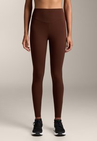 Hnedé legíny s vysokým pásom vyrobené z elastického materiálu, s hladkou textúrou a elegantným dizajnom, v kombinácii s čiernymi športovými topánkami.