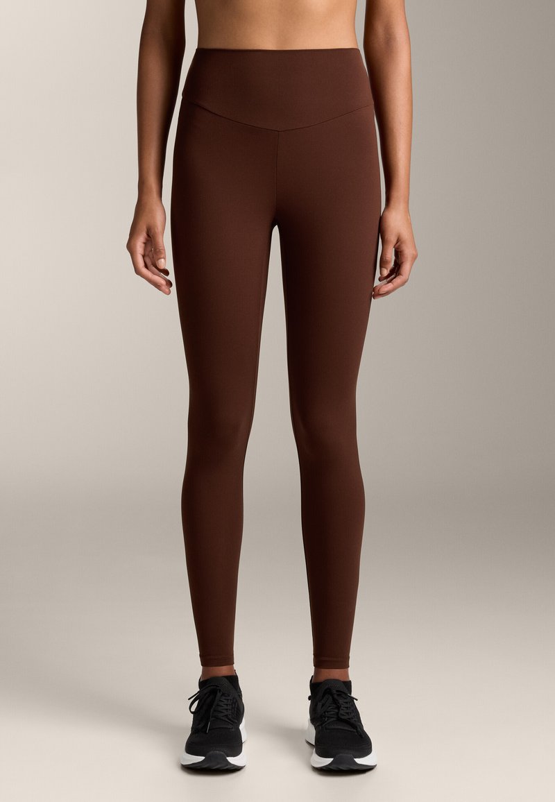 Hnedé legíny s vysokým pásom vyrobené z elastického materiálu, s hladkou textúrou a elegantným dizajnom, v kombinácii s čiernymi športovými topánkami.