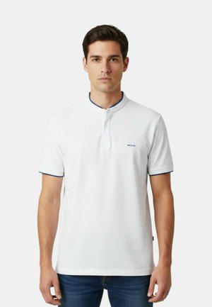 Uomo che indossa una polo bianca a maniche corte con bordatura blu e piccolo logo, abbinata a jeans blu, in piedi davanti a uno sfondo semplice.