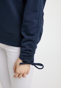 Maglietta a maniche lunghe blu navy con texture arricciata e polsini regolabili. Tessuto morbido, abbinato a pantaloni bianchi attillati.