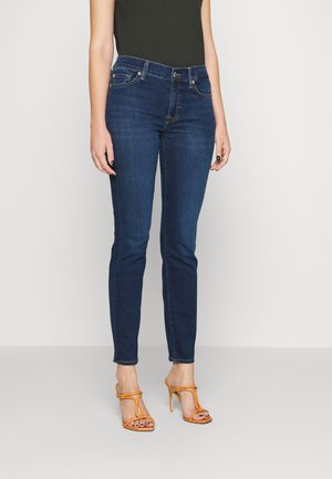 Temno modri skinny jeans z klasičnim pet-pocket dizajnom in subtilnimi zlatimi detajli, v kombinaciji z oranžnimi sandali na visoki peti s trakovi.