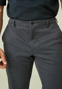 Maßgeschneiderte graue Hose aus strukturiertem Stoff, mit flachem Frontschnitt, Gürtelschlaufen und schwarzem Knopfverschluss. Nahaufnahme der Taille.