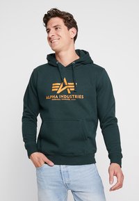 Hoodie verde scuro realizzato in materiale morbido. Presenta una tasca a marsupio e un logo arancione ben visibile, abbinato a un cappuccio con cordino.