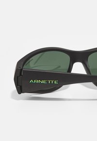 Occhiali da sole neri con lenti verdi. Presentano un audace marchio "ARNETTE" in verde brillante sul braccio. Design elegante e moderno con una finitura opaca.
