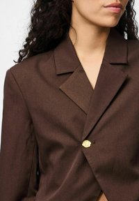 Blazer marron en tissu texturé, avec un col cranté et une fermeture à un seul bouton en or. Coupe ajustée avec un détail de conception en couches.