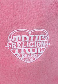 Roze katoenen stof met een wit geborduurd hartlogo met de tekst "TRUE RELIGION BRAND" en decoratieve accenten.