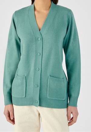 Cardigan boutonné bleu sarcelle clair avec deux poches devant, porté sur un pantalon couleur crème.