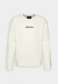 Krémový svetr s dlouhými rukávy, žebrovanými manžetami a lemem. Na hrudi je vyšívané logo "Hollister" v tmavě modré barvě.