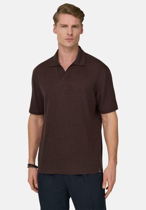 Mann trägt ein dunkelbraunes kurzärmeliges Poloshirt und marineblaue Hose, steht mit neutralem Gesichtsausdruck vor einem einfarbigen Hintergrund.