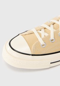 Μπεζ πάνινο αθλητικό παπούτσι με κρεμ καουτσούκ προστατευτικό άκρου και μαύρη επένδυση. Διαθέτει ασημί τελείες για τα κορδόνια και λευκά κορδόνια. Υφασμάτινη υφή με ραμμένα διακοσμητικά στοιχεία.