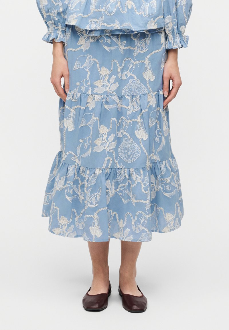 Femme portant une jupe midi bleue et un haut avec des motifs blancs de coquillages et de chaînes, associée à des chaussures plates bordeaux, debout devant un fond blanc.