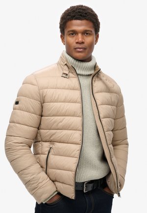 Beige pufferjakke med kviltet tekstur, lynlåslommer og trykknaplukning. Bæres over en lysegrå rullekravesweater.