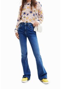 Maglione lavorato a maglia rosa chiaro con motivo floreale, abbinato a jeans a zampa d'elefante blu e sneakers gialli. La modella tiene in mano una pochette bianca.