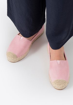 Voeten met roze espadrilles met gevlochten beige neusjes, gecombineerd met losse zwarte broeken, staand op een witte ondergrond.