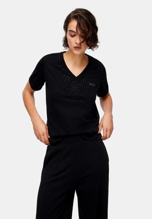 Donna che aggiusta un top nero a maniche corte con scollo a V e piccoli borchiette scintillanti, abbinato a pantaloni neri a gamba larga, su sfondo grigio chiaro.