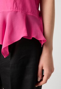 Rosa blus med lager-puffad nederdel, tillverkad av lättviktsmaterial. Blusen är i kombination med svarta, strukturerade byxor. Hand synlig.