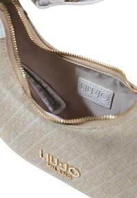 Beige handtas met gestructureerd stoffen patroon, hardware in goudkleur en een ritssluiting. Binnenkant toont een stoffen voering en merklabel.