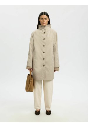 Kvinne iført en beige knappet kåpe, kremfargede bukser, svarte sko, holder en brun semsket veske, stående foran en ensfarget hvit bakgrunn.
