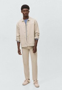 Giacca di jeans beige con bottone frontale, due tasche; abbinata a pantaloni slim-fit beige coordinati e sneakers chiare con dettagli a contrasto.