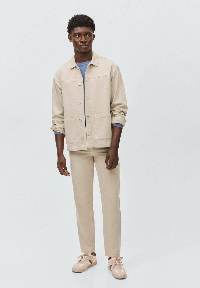 Giacca di jeans beige con bottone frontale, due tasche; abbinata a pantaloni slim-fit beige coordinati e sneakers chiare con dettagli a contrasto.