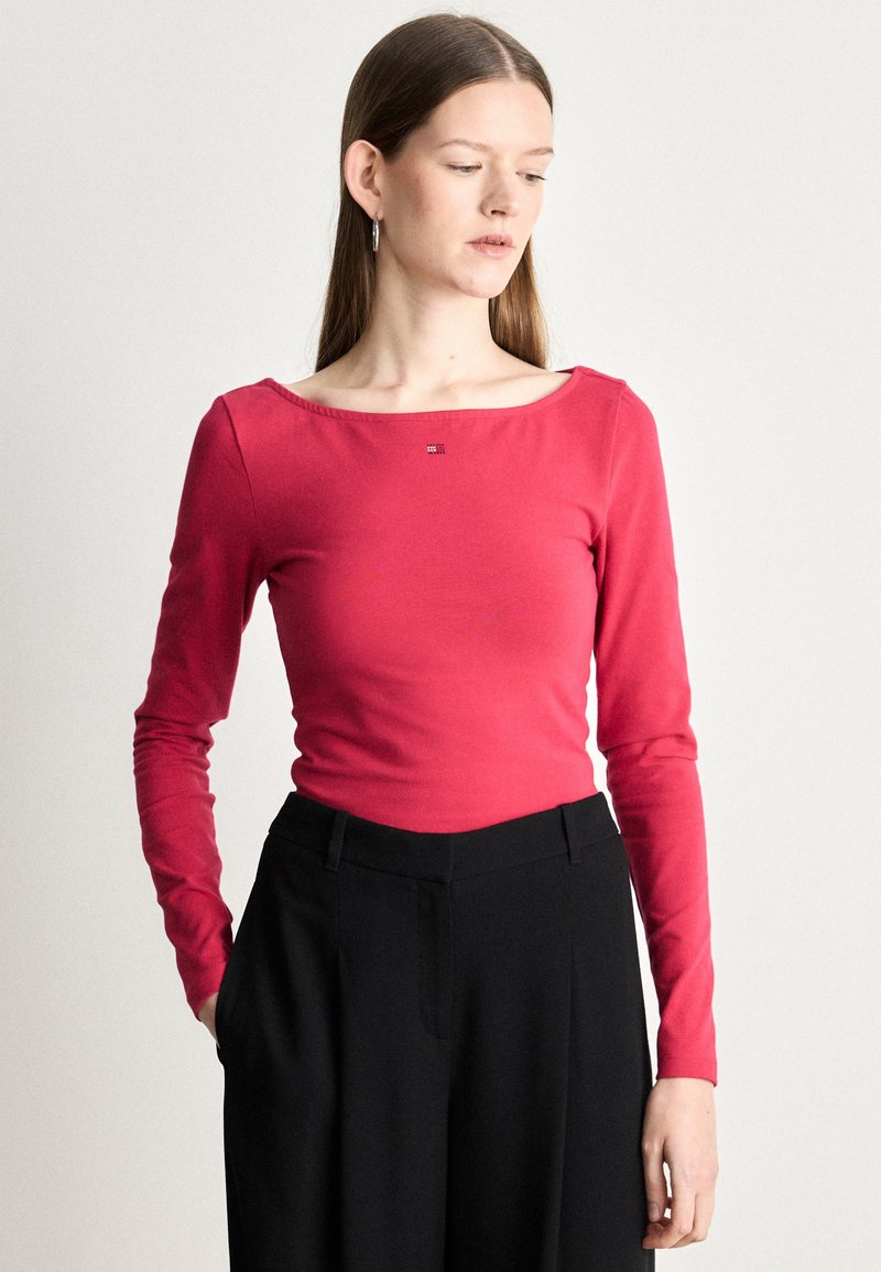 Dolga majica s dolgimi rokavi, prilegajoča v svetlo roza bombažu, z širokim izrezom in subtilnim logotipom. Ujemite jo z visokim pasom in črnimi hlačami.