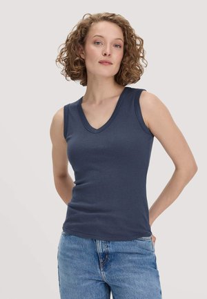 Femme aux cheveux bouclés, debout avec les mains dans le dos, portant un haut sans manches bleu marine à col en V et un jean bleu devant un fond uni.