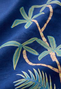 Tissu bleu marine avec un motif de feuilles de palmier en vert et jaune. Les feuilles présentent des contours bien définis. Texture douce avec une finition légèrement brillante.