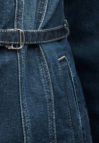 Σκούρο μπλε ύφασμα τζιν με ορατά διπλά ραφές και λεπτή ζώνη τζιν, στερεωμένη με μια μικρή ασημένια αγκράφα.