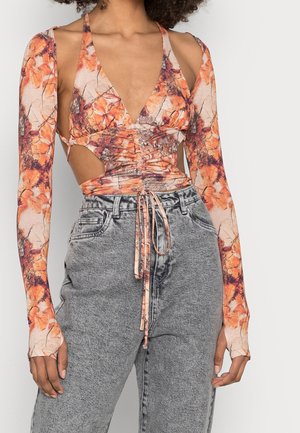 Vrouw draagt een oranje bloemenprint crop top met lange mouwen en uitsparingen, gecombineerd met hoog getailleerde grijze acid-wash jeans.