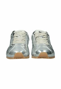 Hopeanväriset metallinhohtoiset tennarit valkoisilla nauhoilla ja kuminruskeilla pohjilla, nähtynä edestä, jossa näkyy brändin tunnisteet kielen päällä.