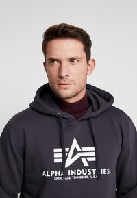 Sudadera gris oscuro hecha de algodón, con un logo y texto en blanco en la parte frontal. Incluye una capucha con cordón y un cuello acanalado.