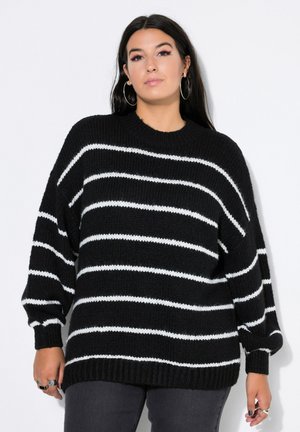 Frau mit langen dunklen Haaren trägt einen schwarz-weißen gestreiften Strickpullover, Ringohrringe und Ringe und steht vor einem schlichten weißen Hintergrund.