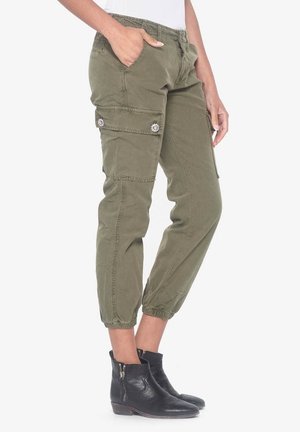 Femme portant un pantalon cargo vert olive avec des poches rabat boutonnées et des bottines noires, debout avec une main dans la poche.