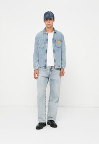Denim jacket in light blue, buttoned, with two chest pockets. Paired with light blue jeans and black shoes. Accessories include a blue cap.
