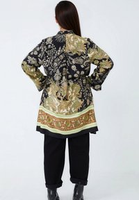 Zwarte, gestructureerde lange jas met gouden en groene bloemmotieven, met een riem in de taille. Gecombineerd met losse zwarte broeken en ankle boots.