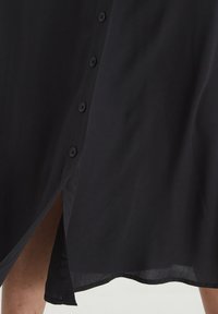 Černé košilové šaty s knoflíky a bočním rozparkem, vyrobené z lehké látky. Mají šest sladěných knoflíků a hladký, mírně texturovaný povrch.