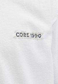 Λευκό ύφασμα με κεντημένο κείμενο "CORE 1990" σε μαύρο και αχνή καλλιγραφική γραφή σε ανοιχτό γκρι πίσω από αυτό.